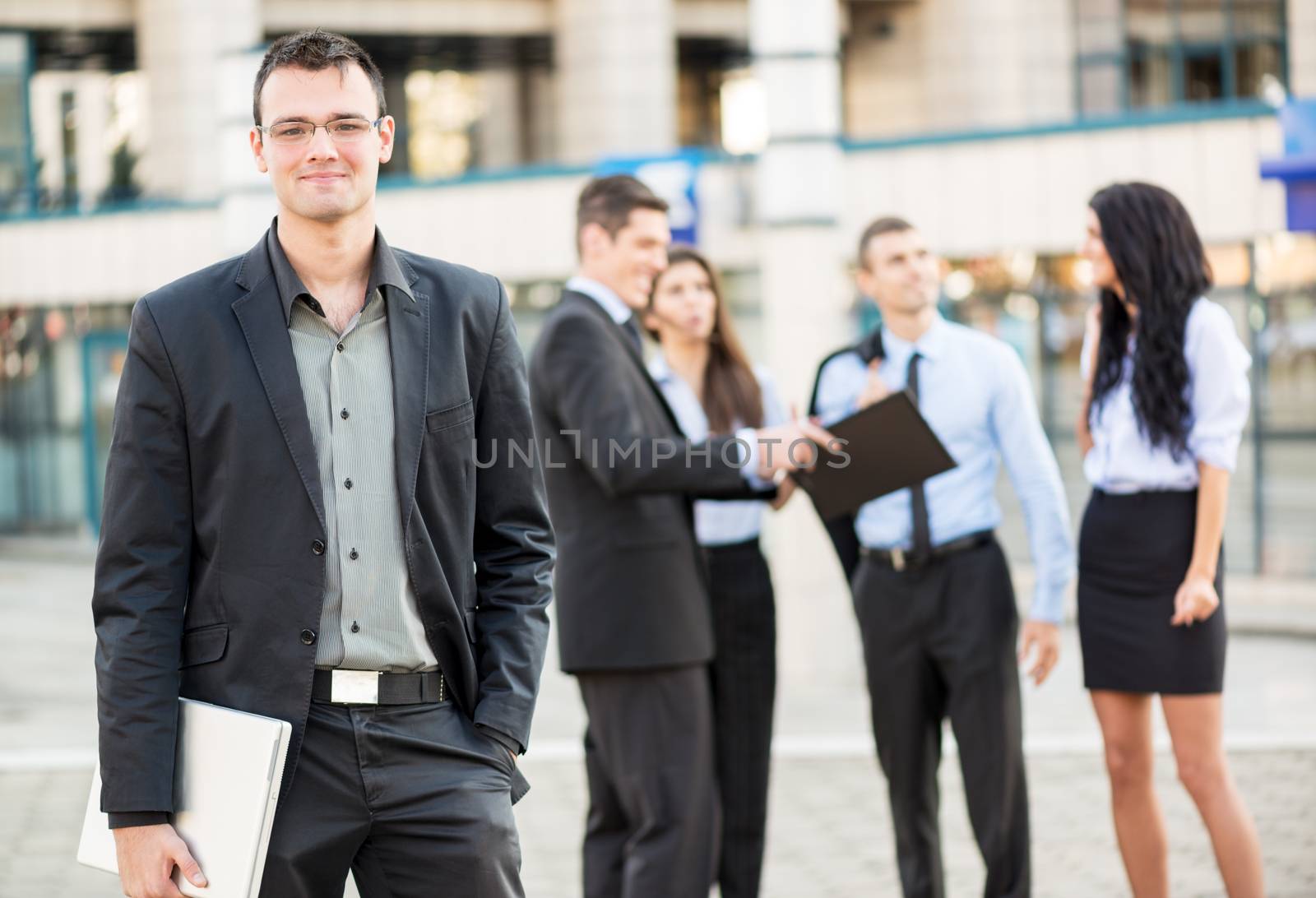 Young businessman with a laptop standing in front of office building separated from the rest of the business team. With a smile looking at the camera.