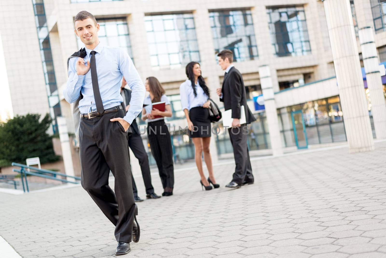 Young smiling businessman standing in front of office building separated from the rest of the business team.