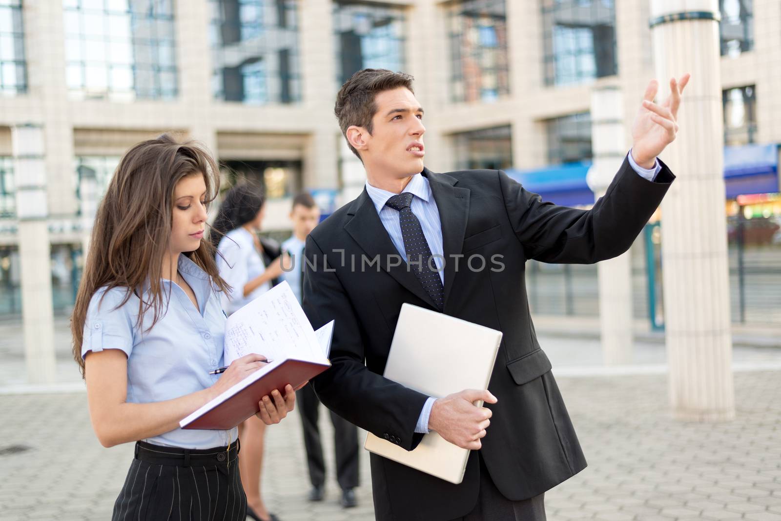 Young businessman and his secretary standing in front of office building.  Businessman carrying in one hand a closed laptop while his other hand pointing to the heights, and the secretary keeps the planner.