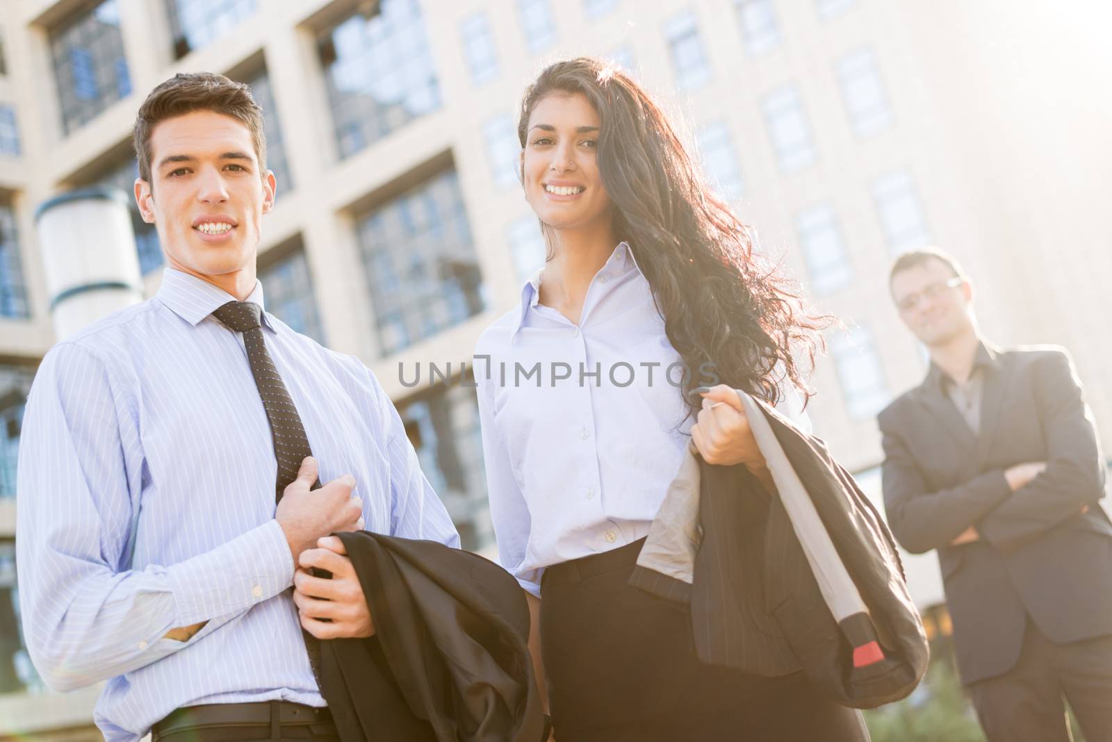 Young businesswoman and businessman standing in front of office building, with a smile looking at camera,  while from behind the sun shines.