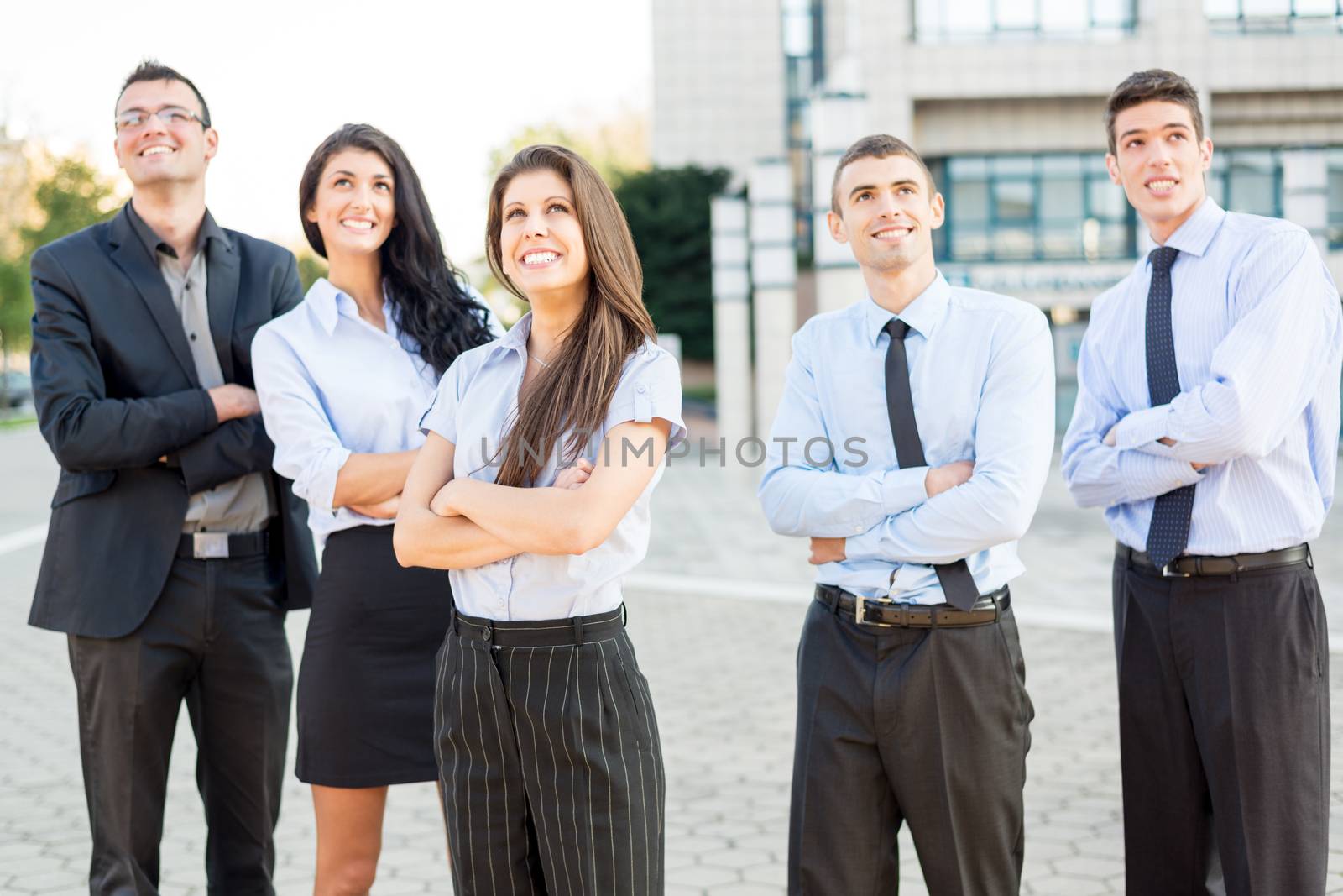 A small group of young business people standing in front of office building with arms crossed and with a smile on their faces looking in height.