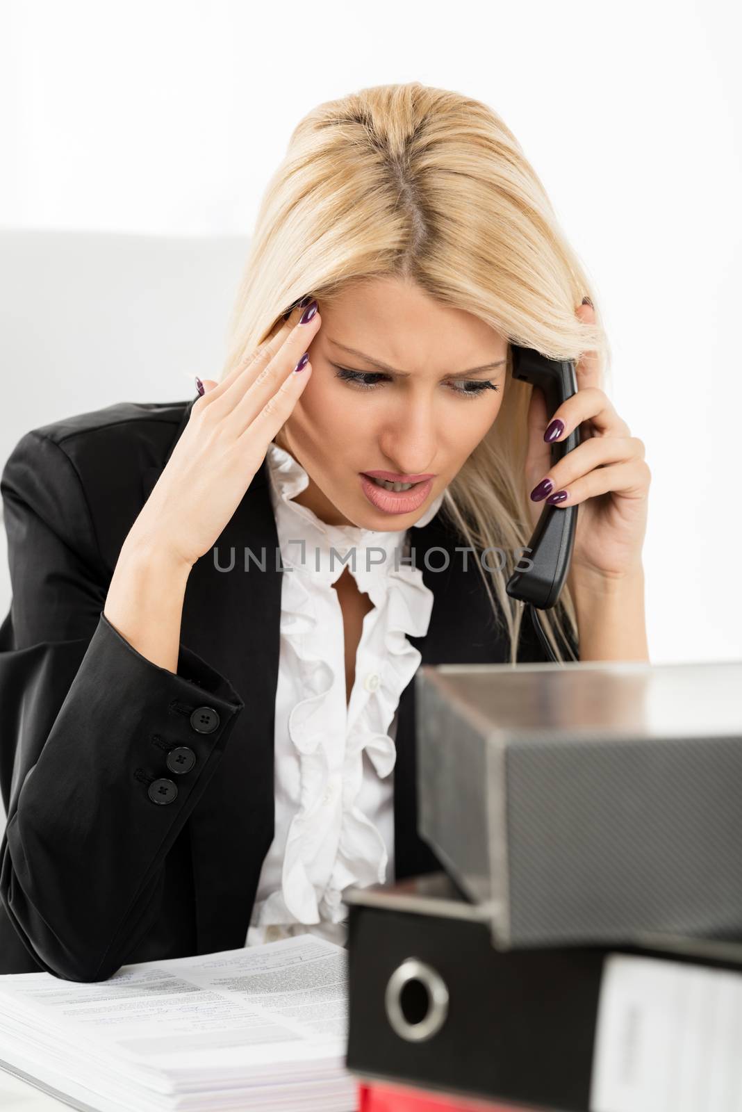 Young businesswoman, phoning at the office, with a worried expression on her face. In front of her is a bunch of binders.