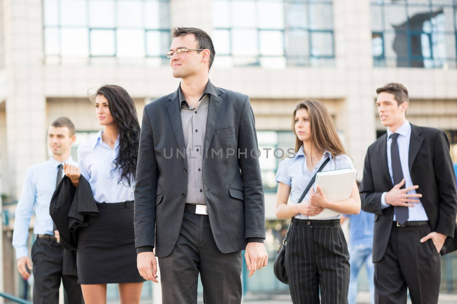 Young businessman, elegantly dressed, standing  with his team of young businesswomen and businessmen in front of office building. Everyone is looking to the side.