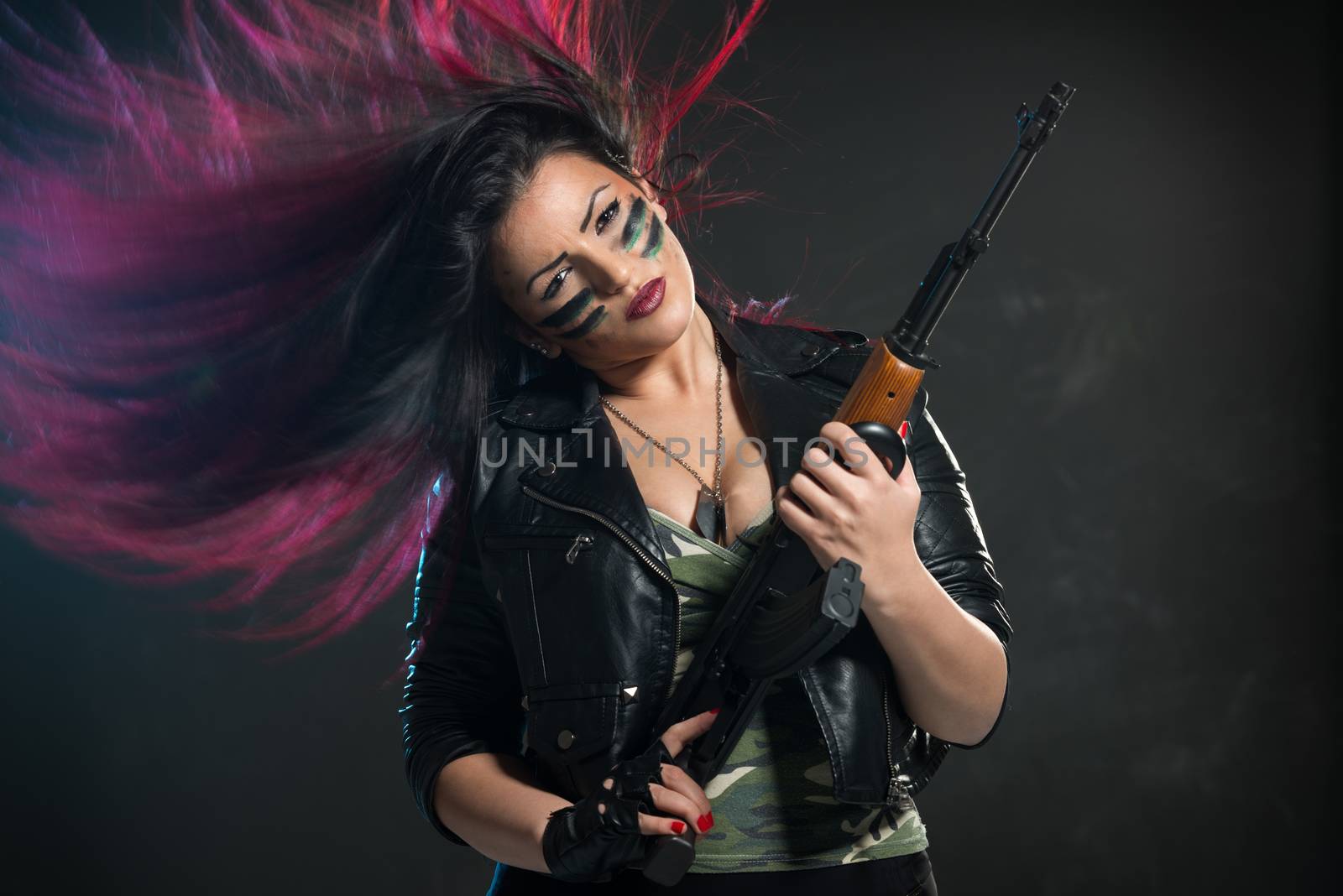 Attractive young woman with disheveled dyed hair is holding an automatic rifle and dangerous expression on her face, painted war paint, looking at the camera.