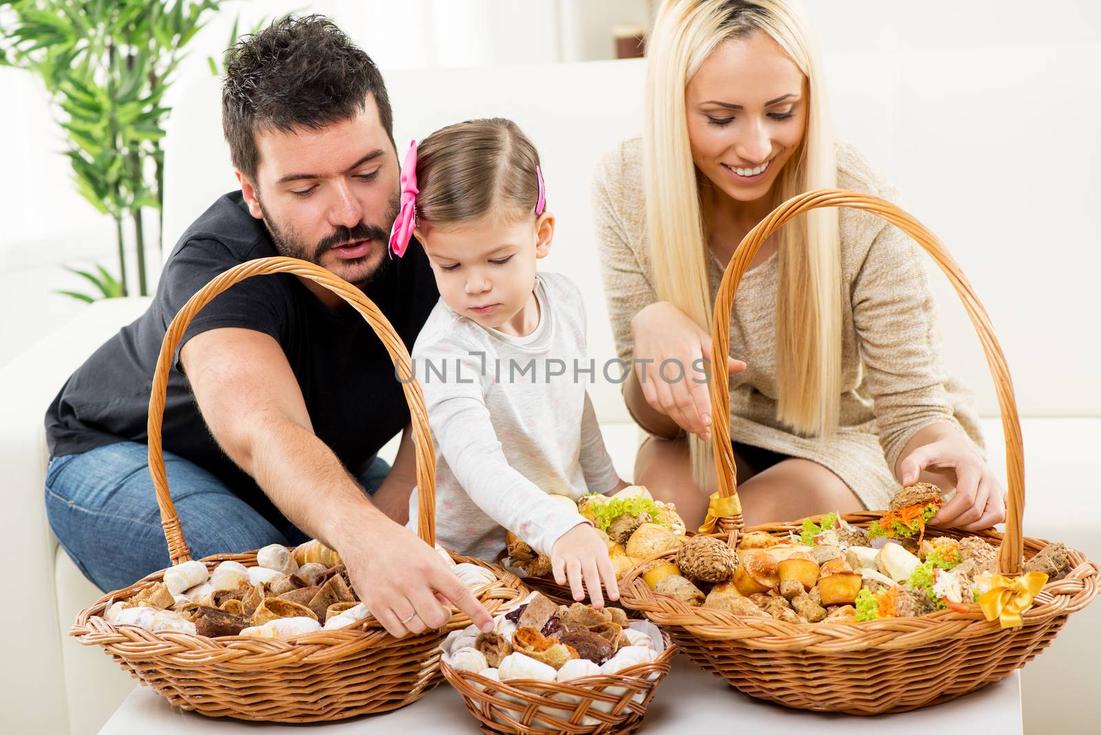 Happy family together, parents with daughter sitting on the couch in the living room in front of woven baskets filled with pastry, choosing the next snack.