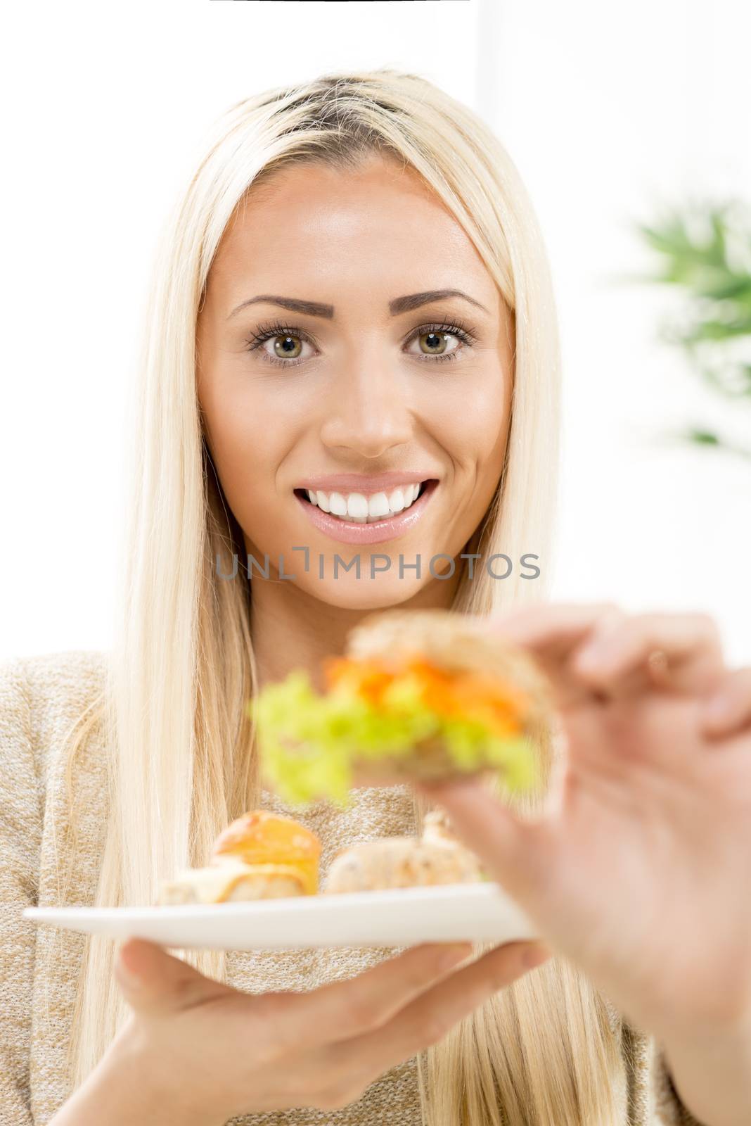 Beautiful young blonde woman with a smile on her face looking at the camera, holding a mini sandwich that is out of focus, offering the viewer a bit.