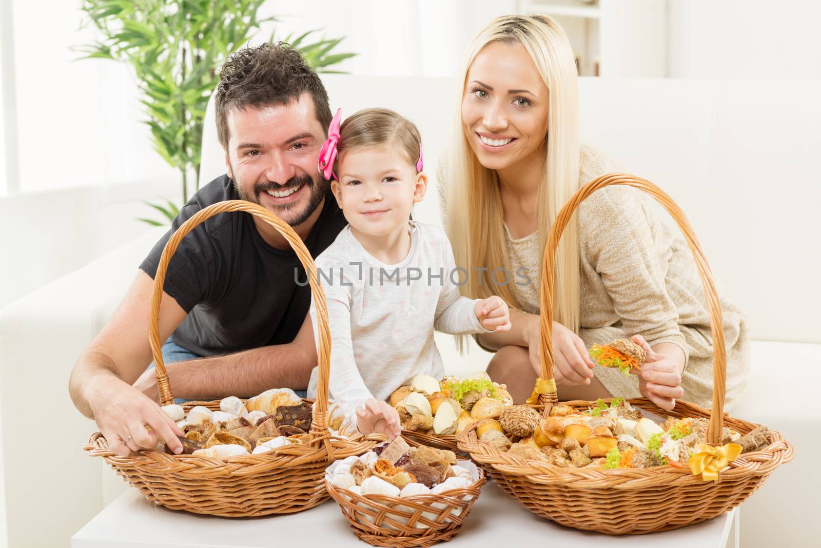Happy family together, parents with daughter sitting on the couch in the living room in front of woven baskets filled with pastry.