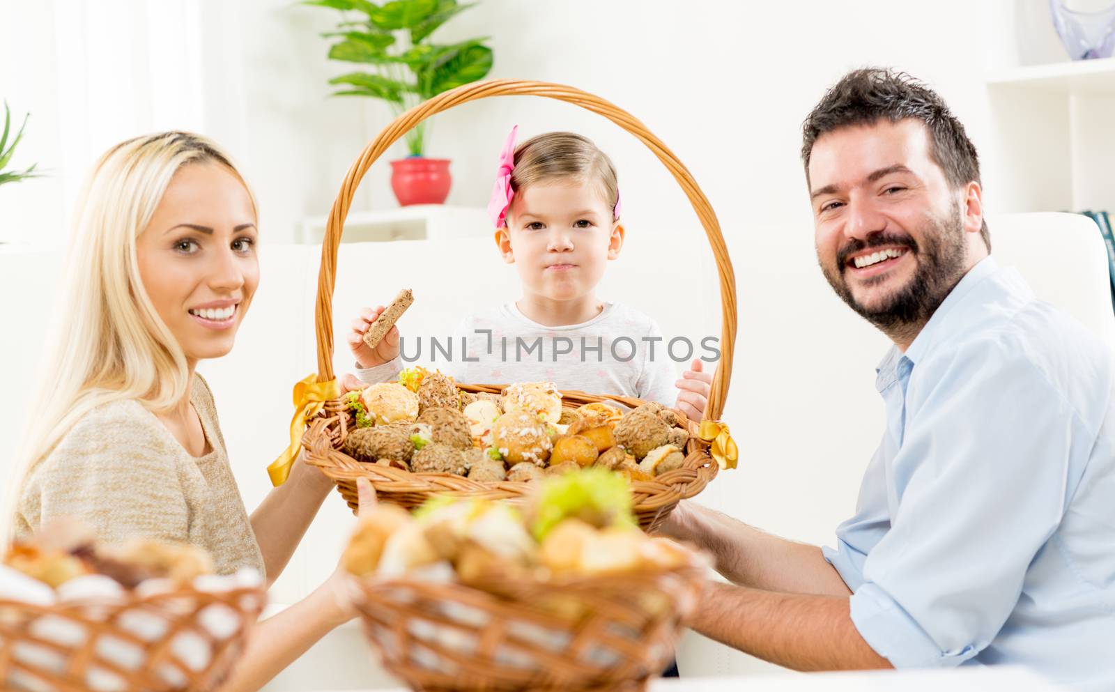 Young parents kneel in front of their little daughter, holding large woven basket with pastries, and with a smile looking at the camera.