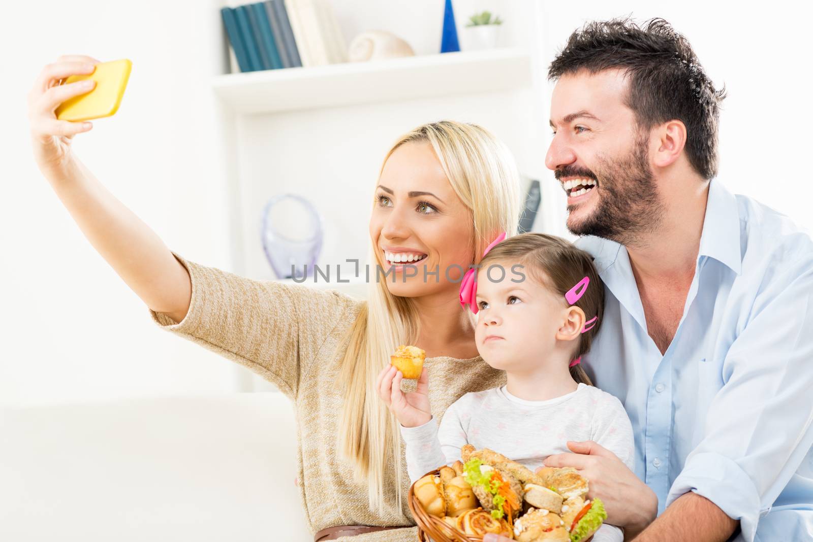 Young beautiful woman photographed with a mobile phone herself, her husband and their beautiful daughter with a wicker basket filled with pastry products.