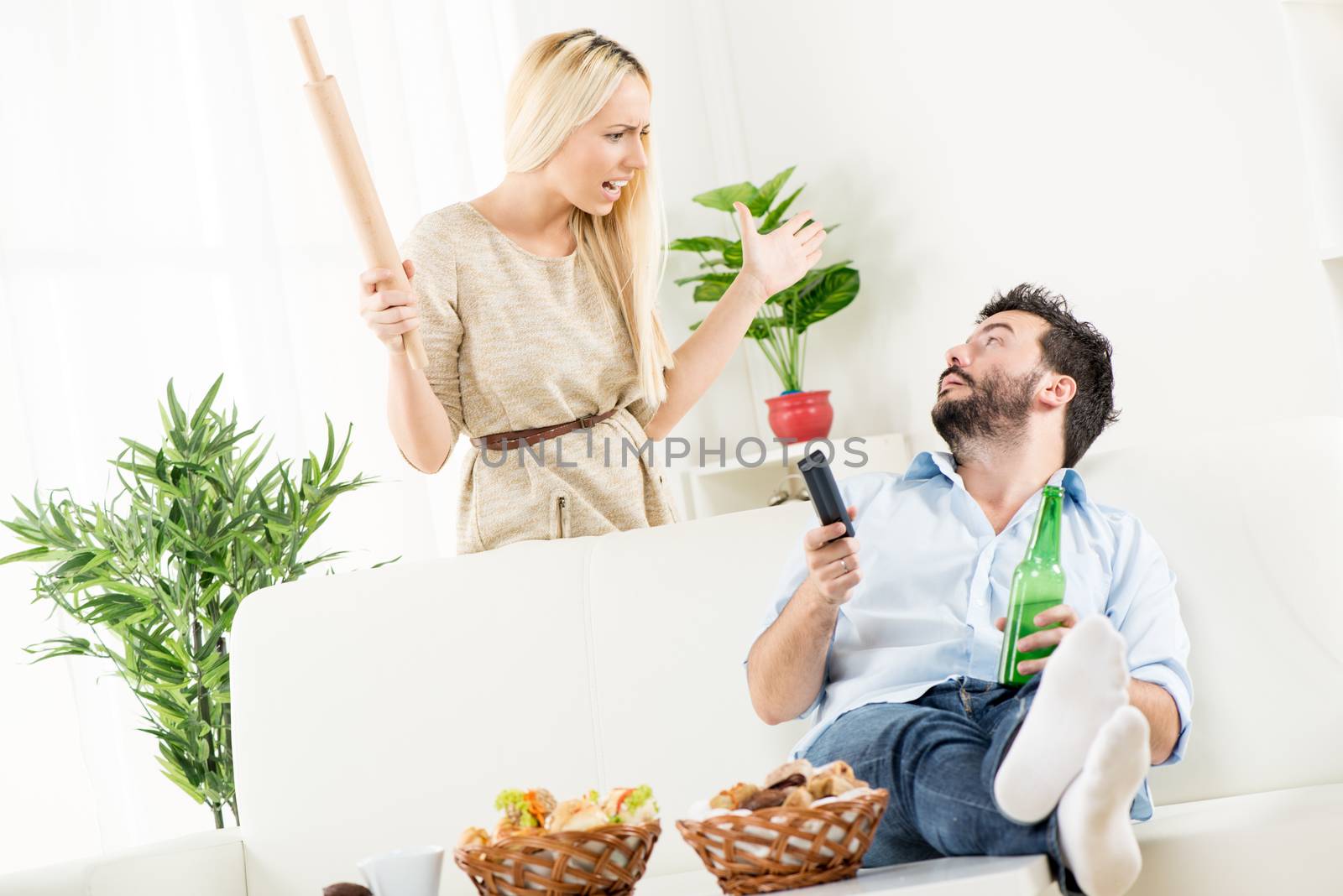 Young man sitting on a couch next to a basket of pastries with his feet up on the table, holding a bottle of beer in one hand, remote control in the other hand,looks at the woman standing behind him with a rolling pin in her hand and an angry expression on her face.