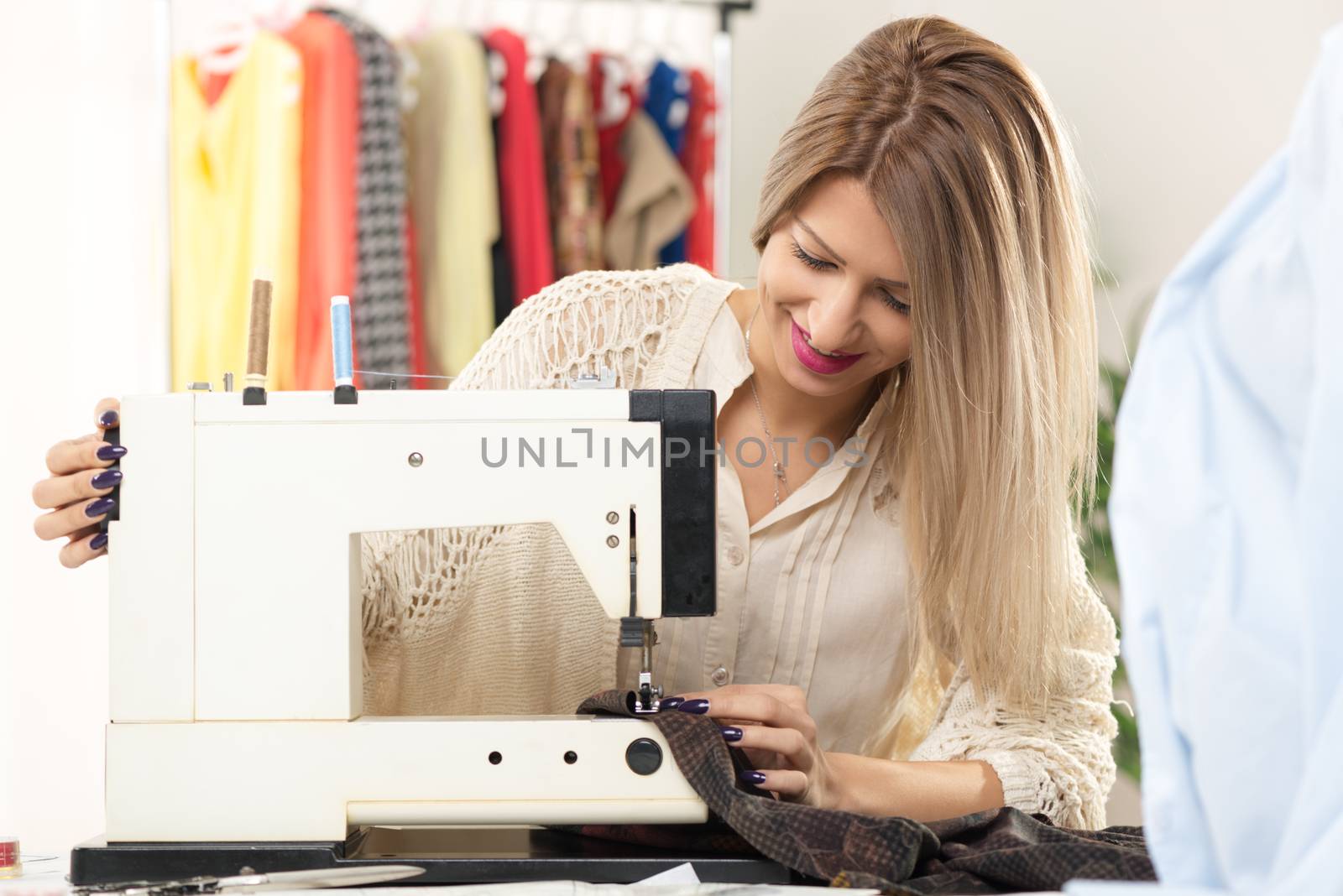 Young beautiful smiling girl sew with a sewing machine, in the background you can see hanging garments.