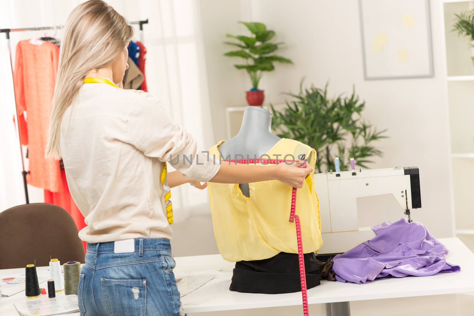 Young women, photographed from behind, with a tape measure, takes measure of clothing which standing on a mannequin. Beside her on the table there is a sewing machine.