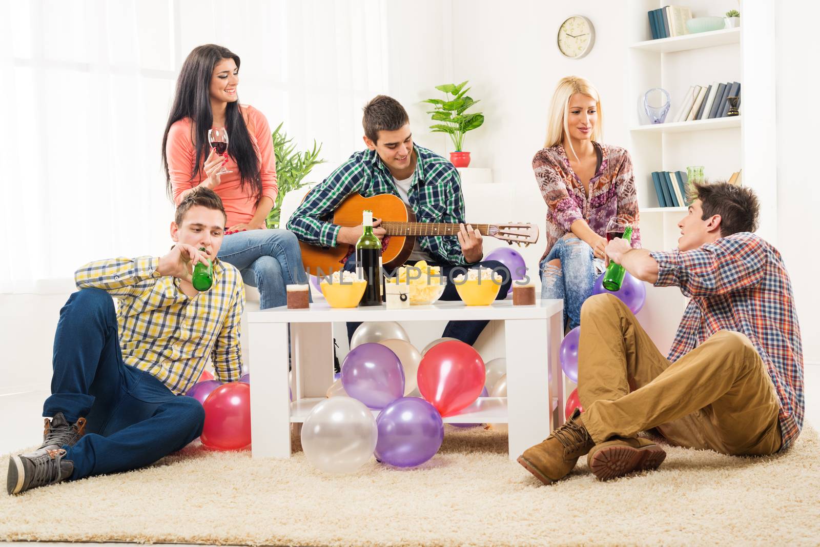 Small group of young people at the home party, enjoy the sounds of the acoustic guitar, drinking wine and beer.