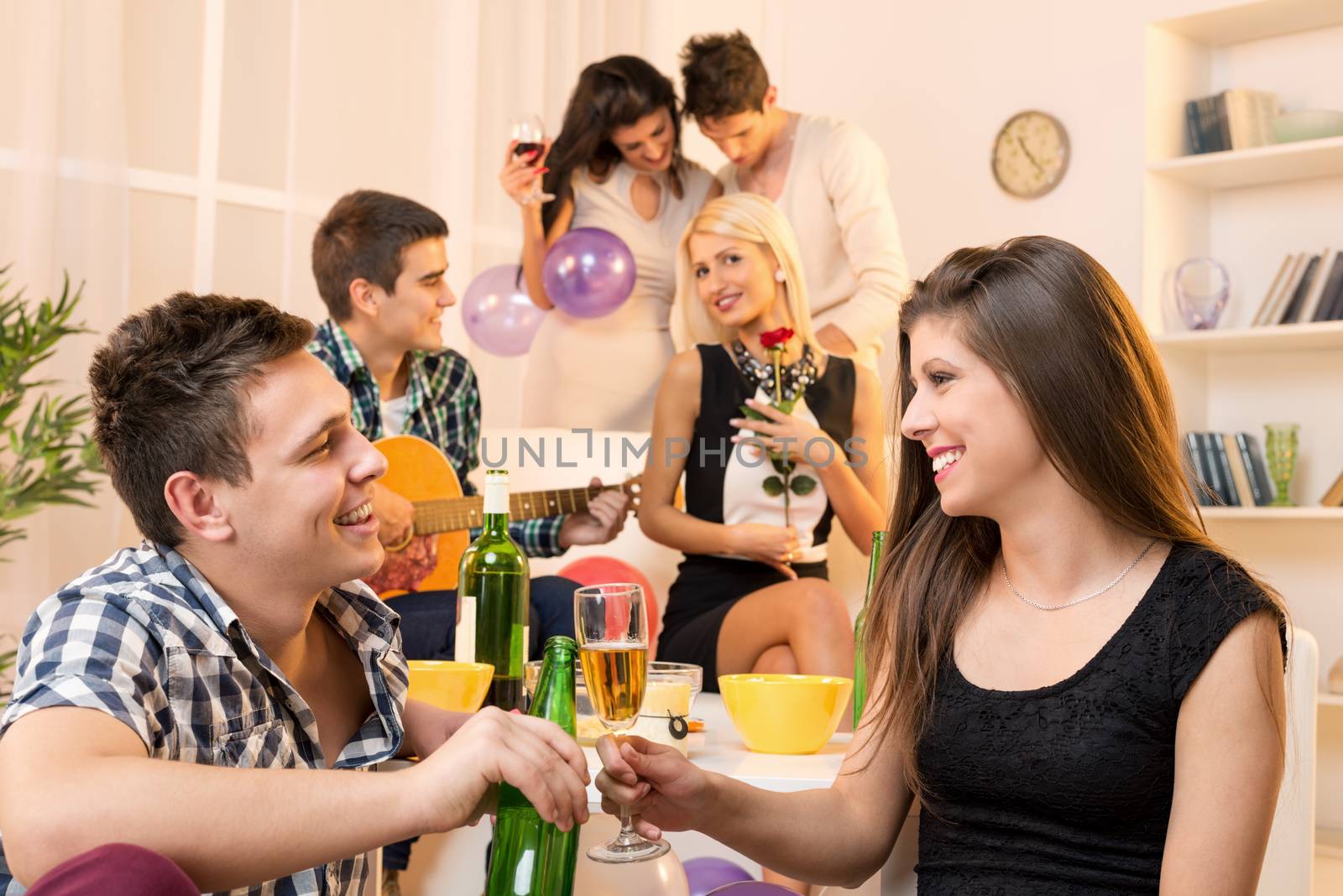 Young couple at a house party, sit on the floor and knocking with drinks, in the background you can see their friends, two couples.