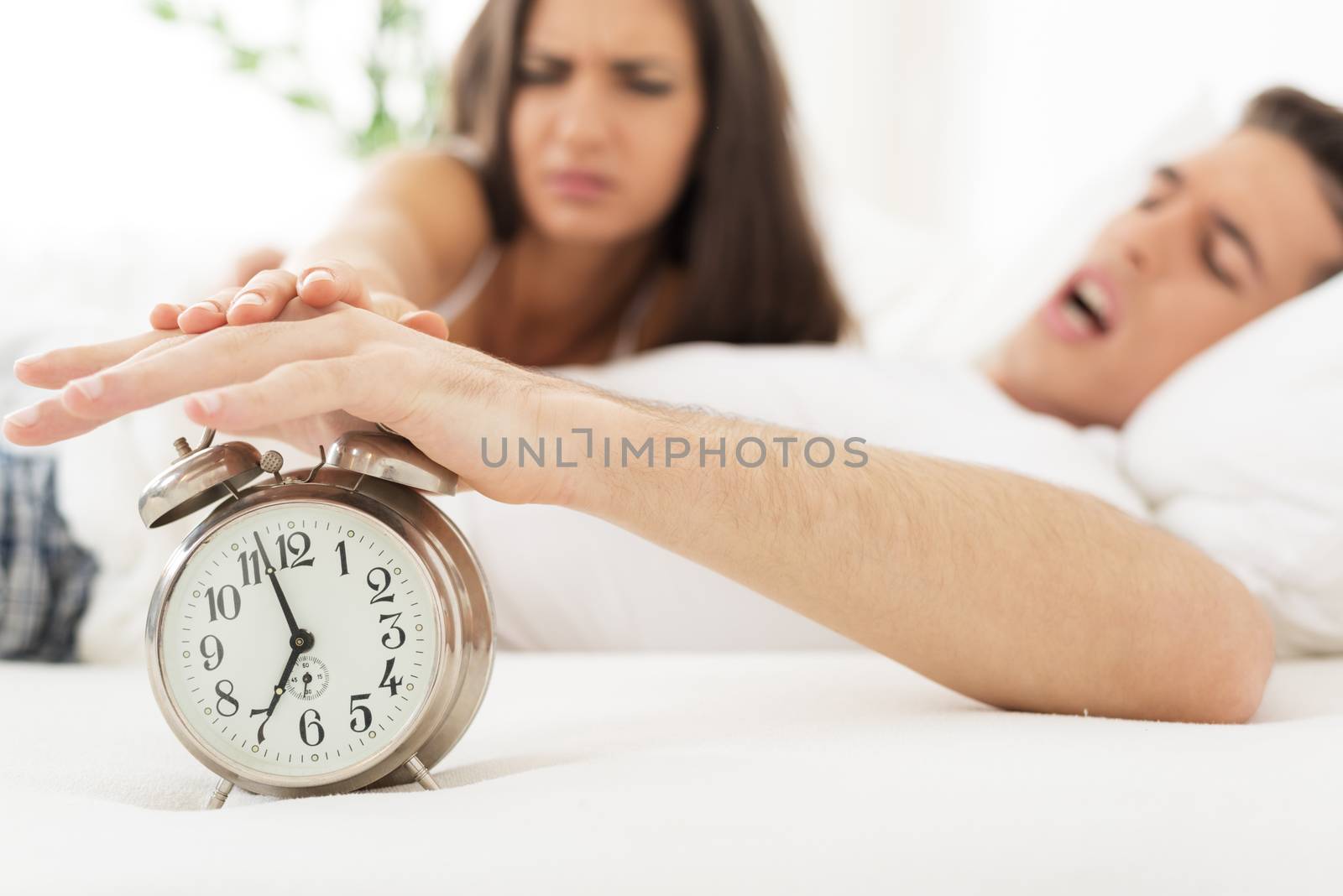 Young couple in bed with an expression of anger on their faces, awaken to the ringing of the alarm clock, the hands reaching toward him, to turn it off. Alarm clock is in the foreground.