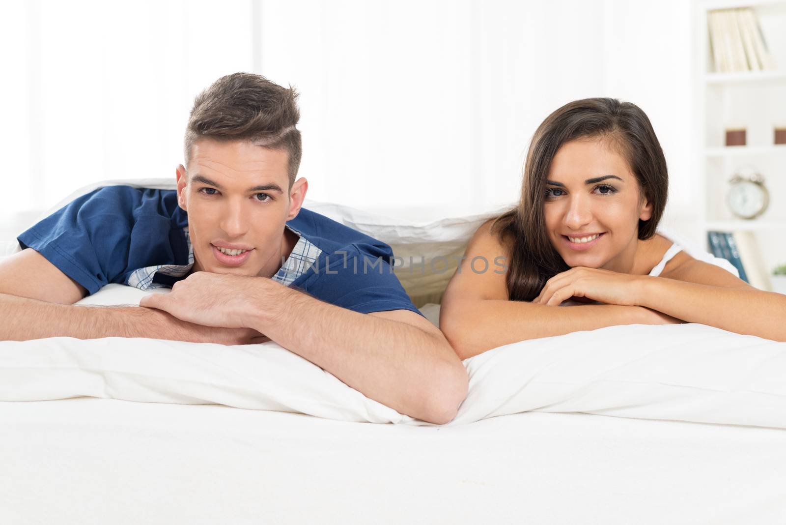 Young heterosexual couple in bed, lying under the duvet with a smile looking at the camera.