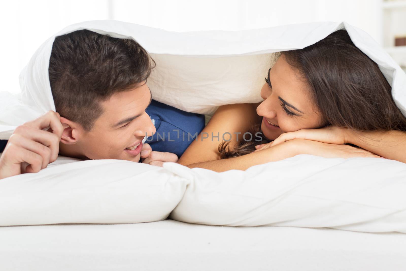 Young heterosexual couple in bed, lying under the duvet with a smile looking at each other.