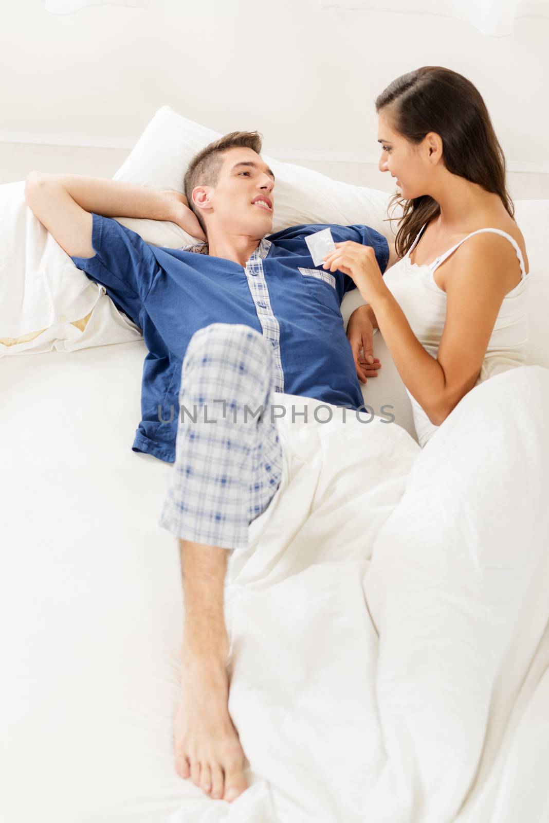 Young heterosexual couple in bed. Girl holding a condom in her hand and looking at a guy in pajamas who lying next to her in bed.