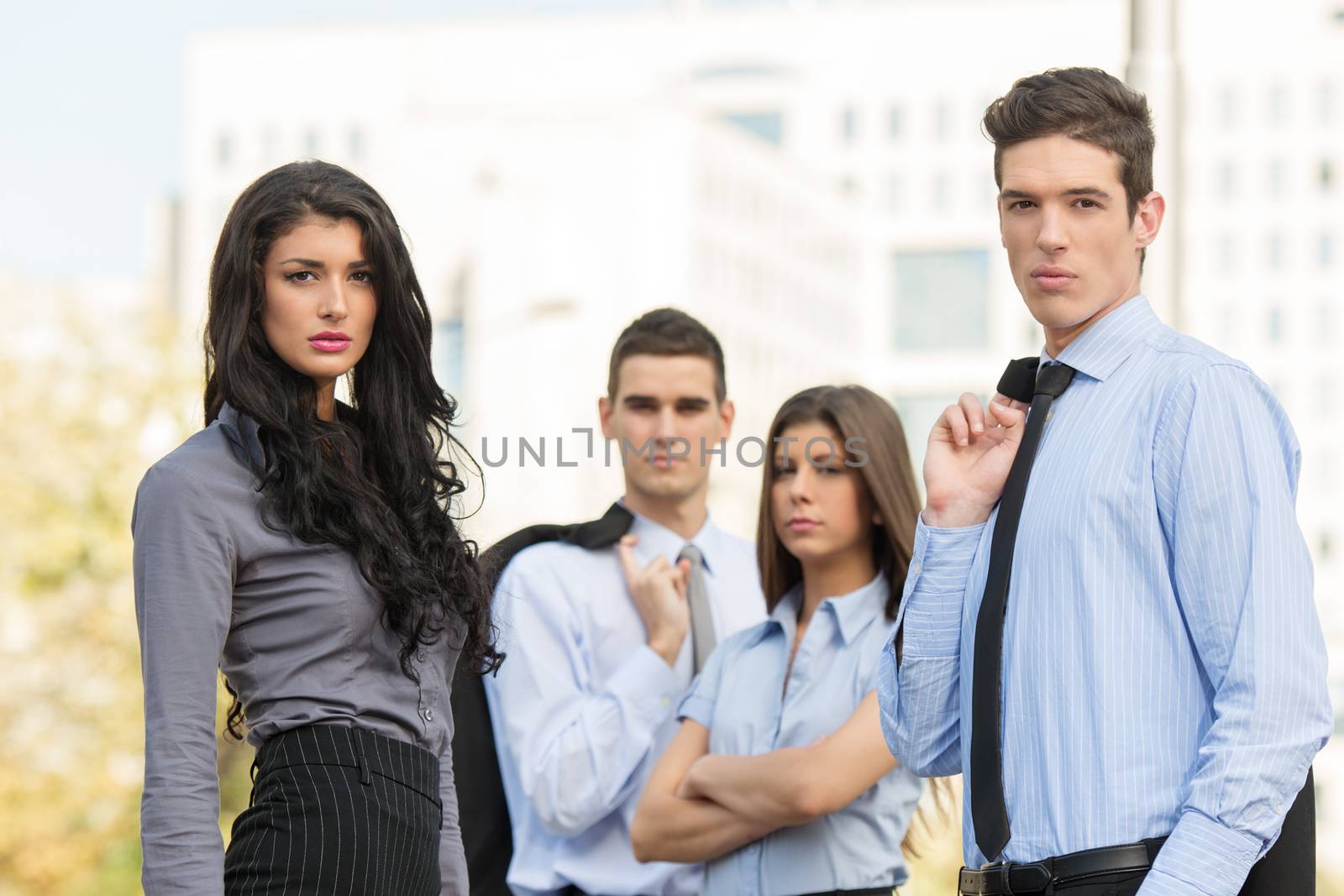 Two couples of young handsome business people, elegantly dressed, standing outside, with a serious expression on their faces looking at the camera. In the background looms large office building.