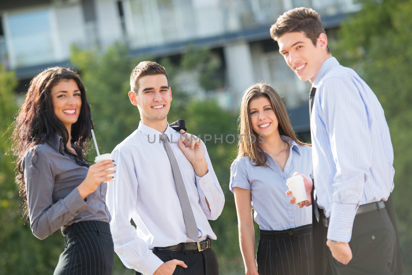 Group of young business people standing outside near their company on the coffee break, enjoying the beautiful day and with a smile on their faces looking at the camera.