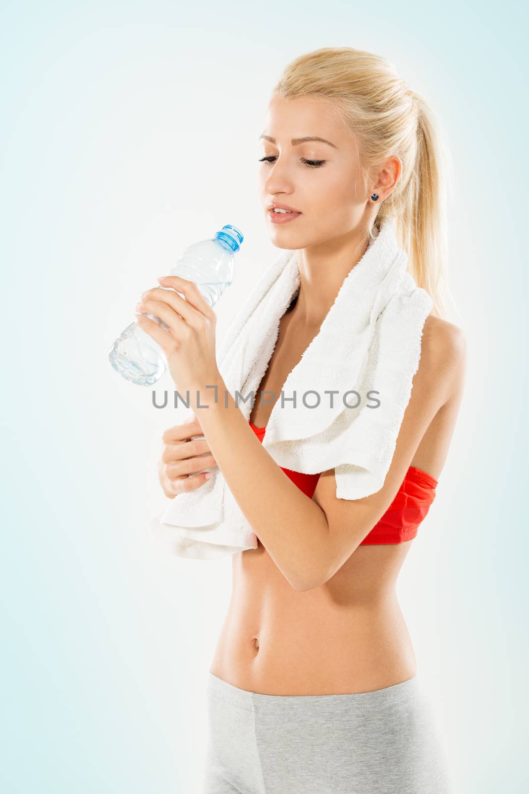 Beautiful blonde girl in sports clothes with a towel over her shoulders, drinking water from a bottle.