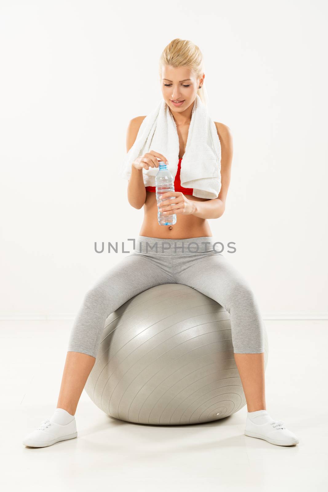 Young pretty blonde girl in sports clothes with a towel over her shoulders and a bottle of water in hand, sitting on the fitness ball.