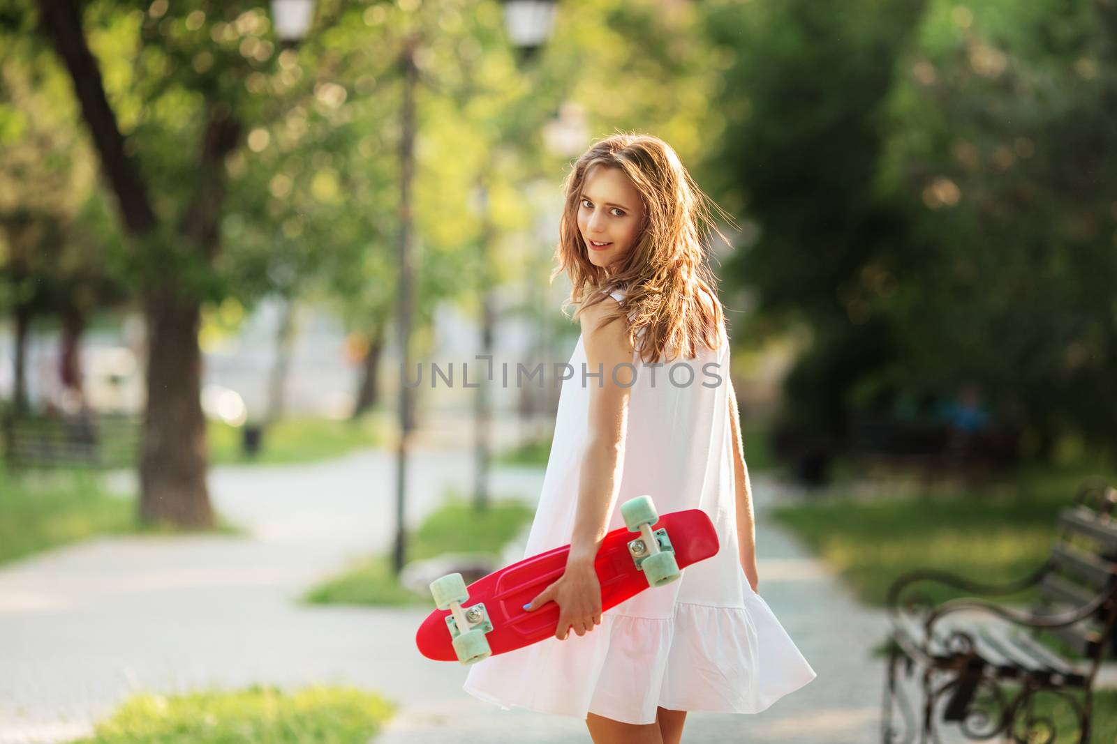 Portrait of lovely urban girl in white dress with a pink skateboard. Portrait of a happy smiling woman. Girl standing with a pink skateboard outdoors. Girl rides on the asphalt on a plastic skateboard