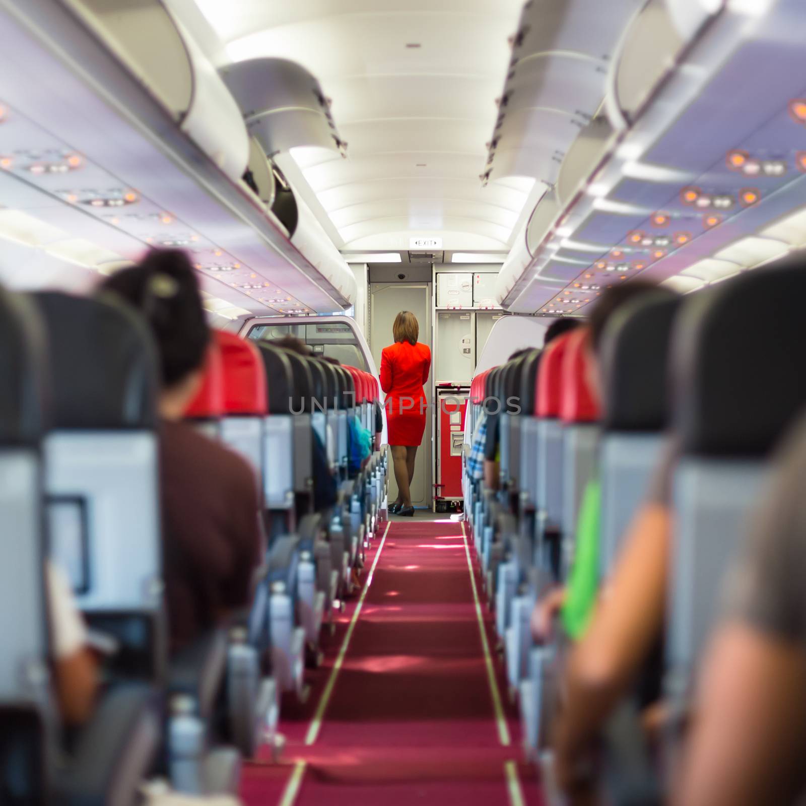 Interior of airplane with passengers on seats and stewardess in red uniform at the aisle. 