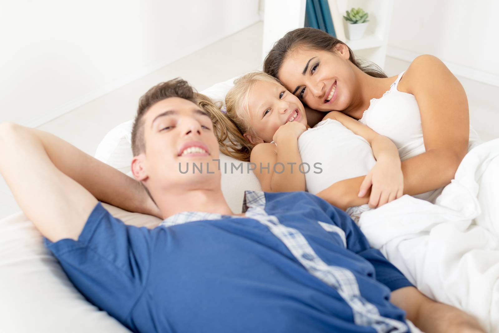 Young parents with their little daughter between them, lie in bed and with a smile looking at camera.