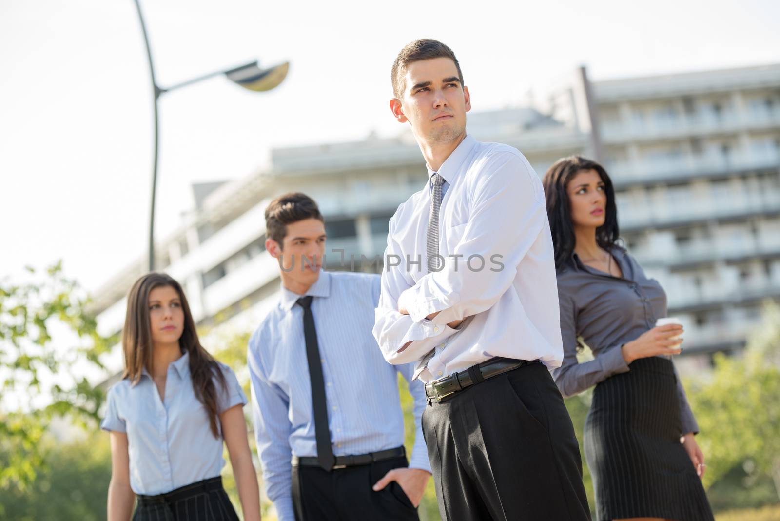 Young businessman standing outside near the office building separated from the rest of the business team  and   looking into the distance with a serious expression on his face.