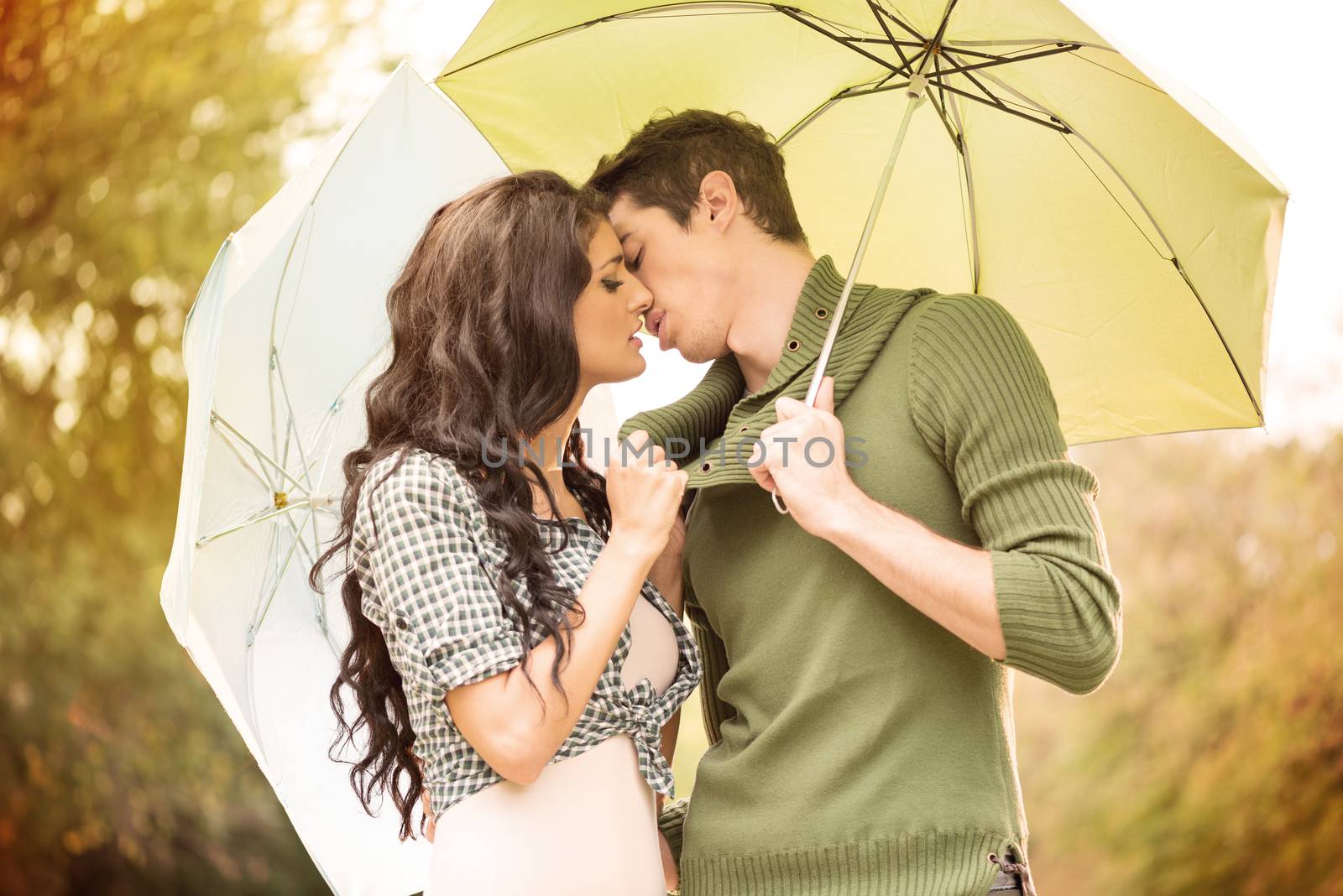 Young girl  and a boy standing embracing under umbrellas and they kiss. In the background is greenery.