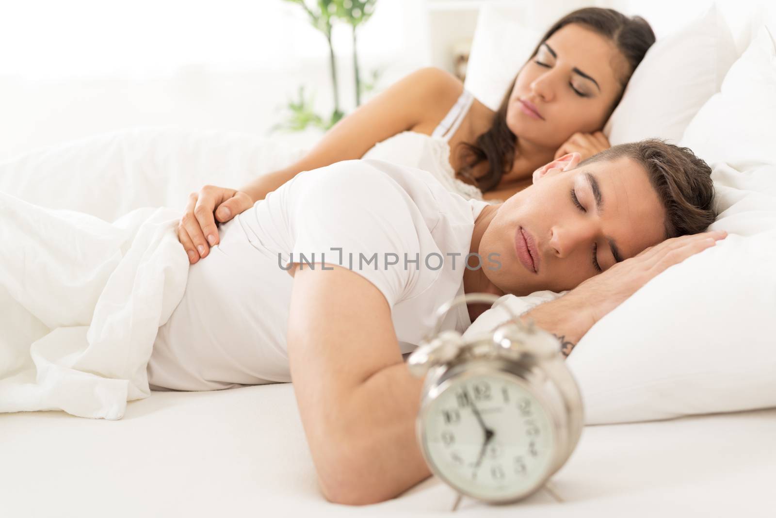 Young heterosexual couple sleeping in bed next to an alarm clock.