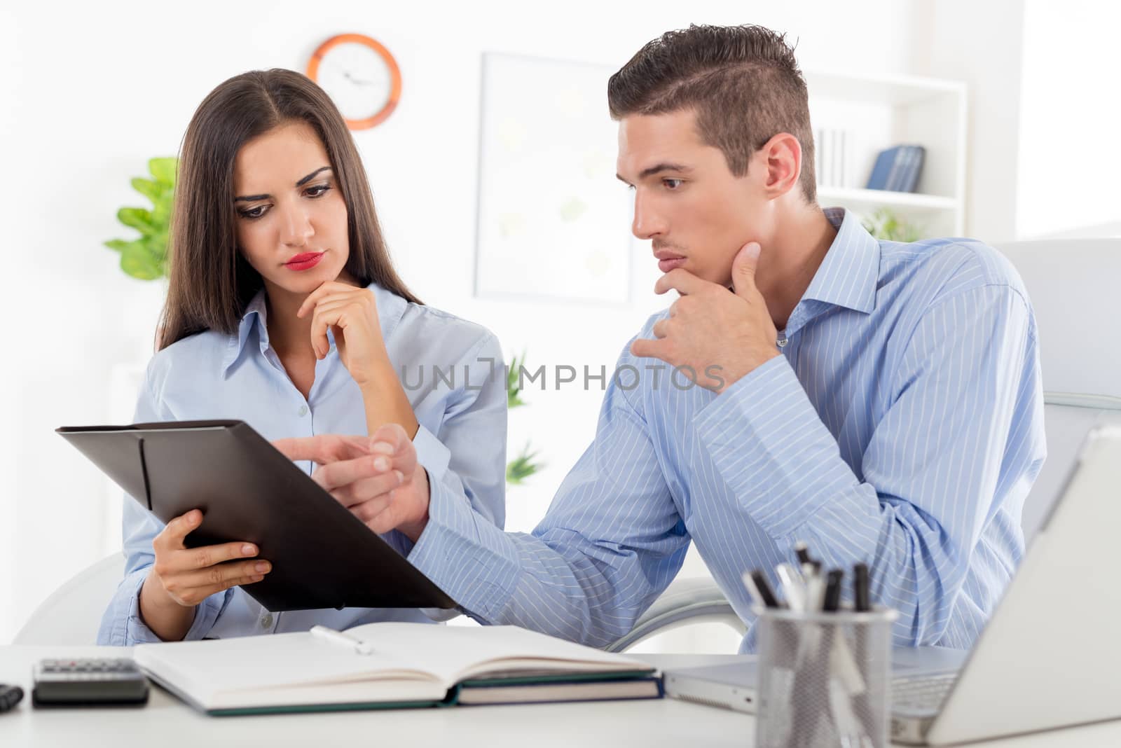 Young businesswoman and businessman, sitting in the office at an office desk with a serious expression on their faces look in the folder that holds a business woman.