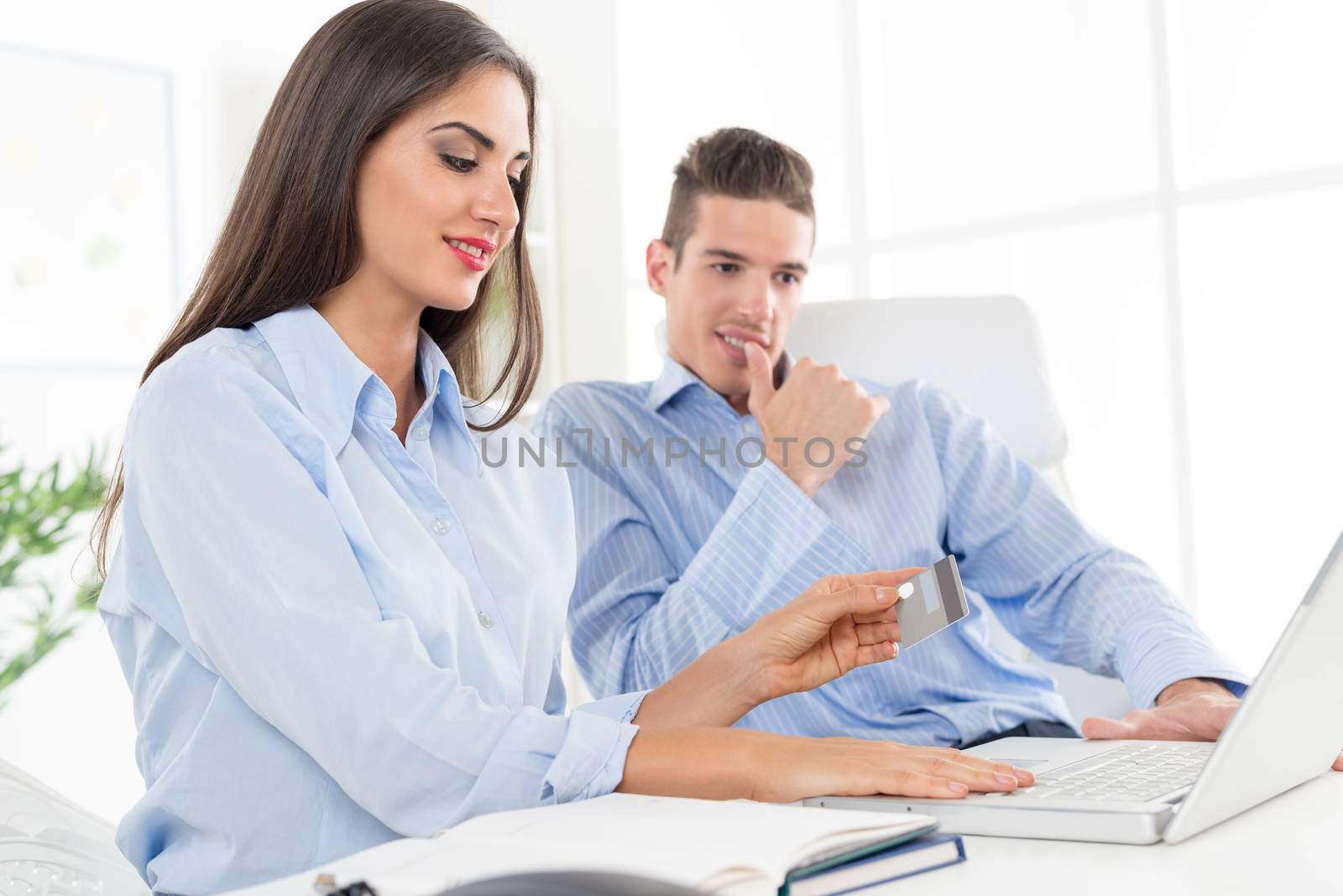 Two young smiling business people, man and woman sitting in office at an office desk in front of a laptop. Woman holding a credit card.