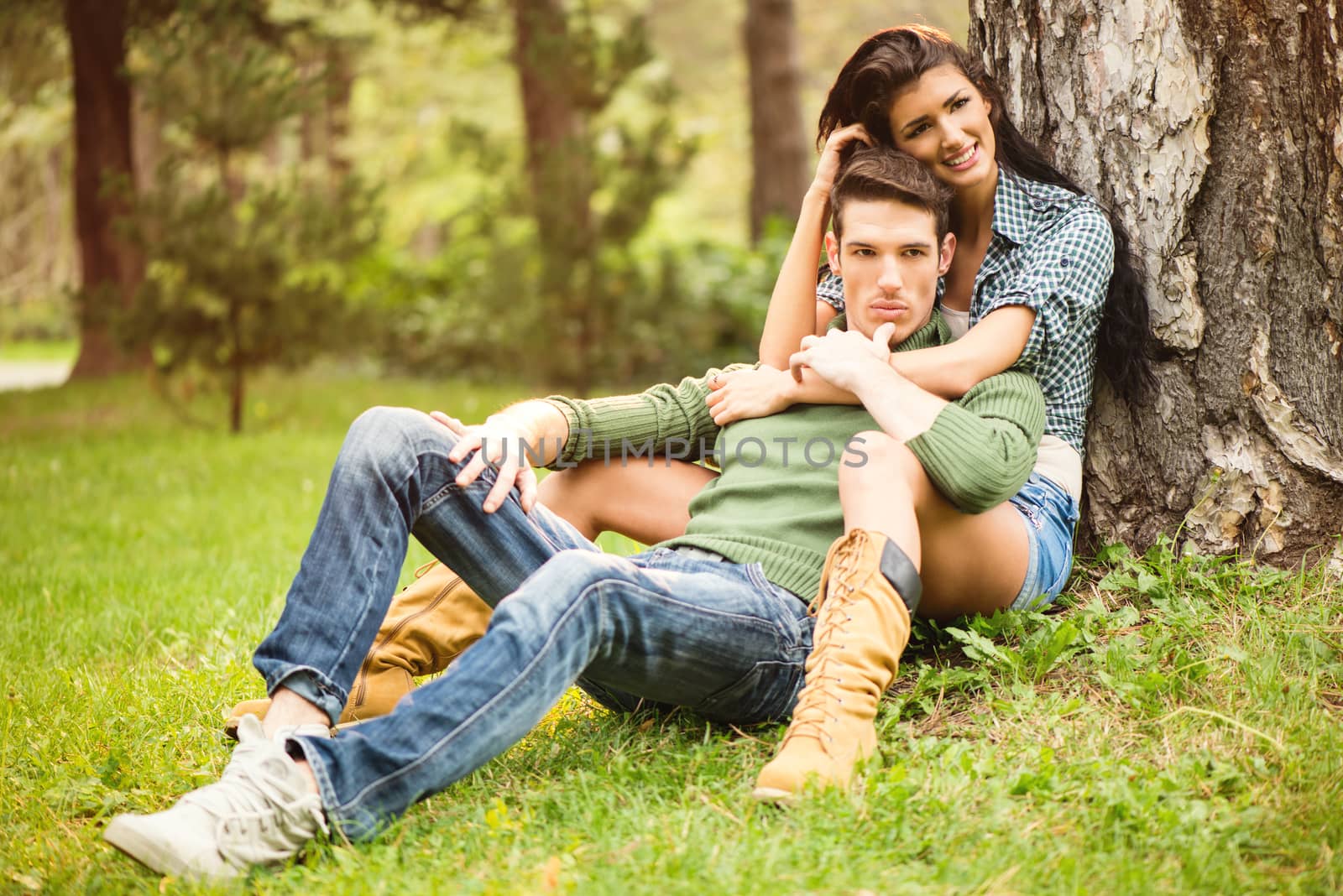 Beautiful brunette girl with a handsome guy, sitting on the grass in the park. The girl was leaning against a tree with a smile looking at the camera, a guy sitting among the girl's legs, leaning back on her.
