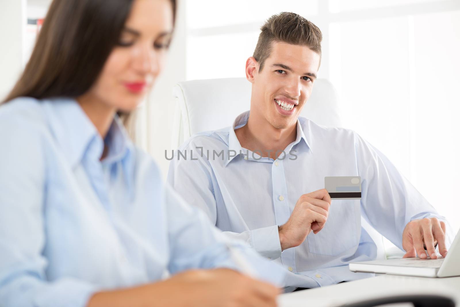 Young business people, man and woman, sitting in an office, a young businessman who is in the foreground holding a credit card and with a smile looking at the camera.