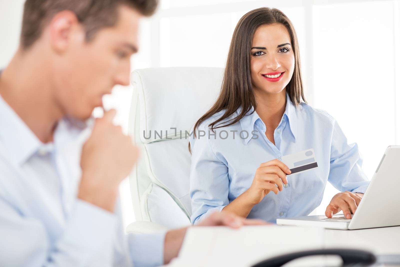 Young business people, woman and man, sitting in an office, a young businesswoman which is in the foreground holding a credit card and with a smile looking at the camera.