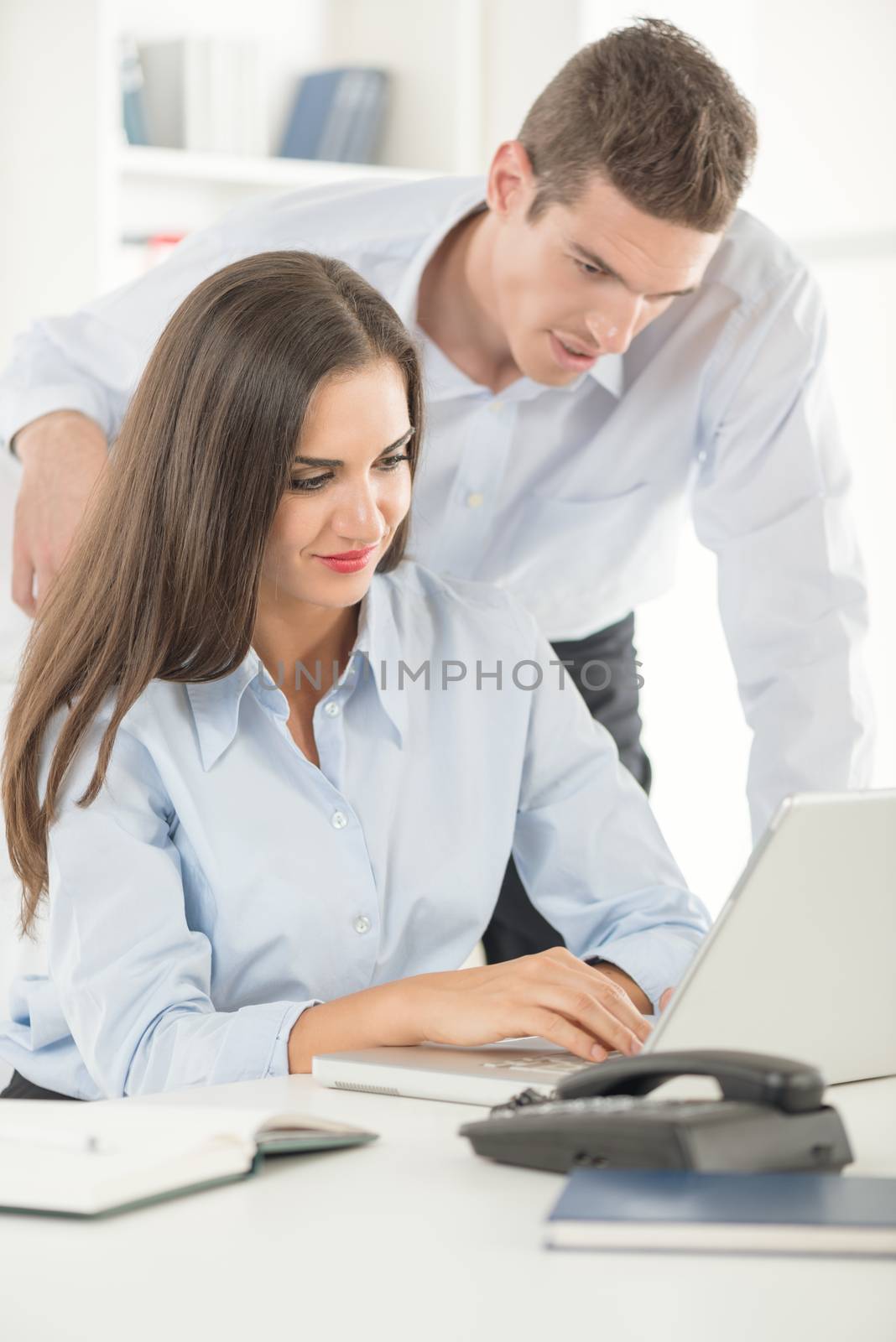 Two young business people in office, woman sitting at an office desk and typing on laptop, behind her standing a young businessman and looking at laptop.