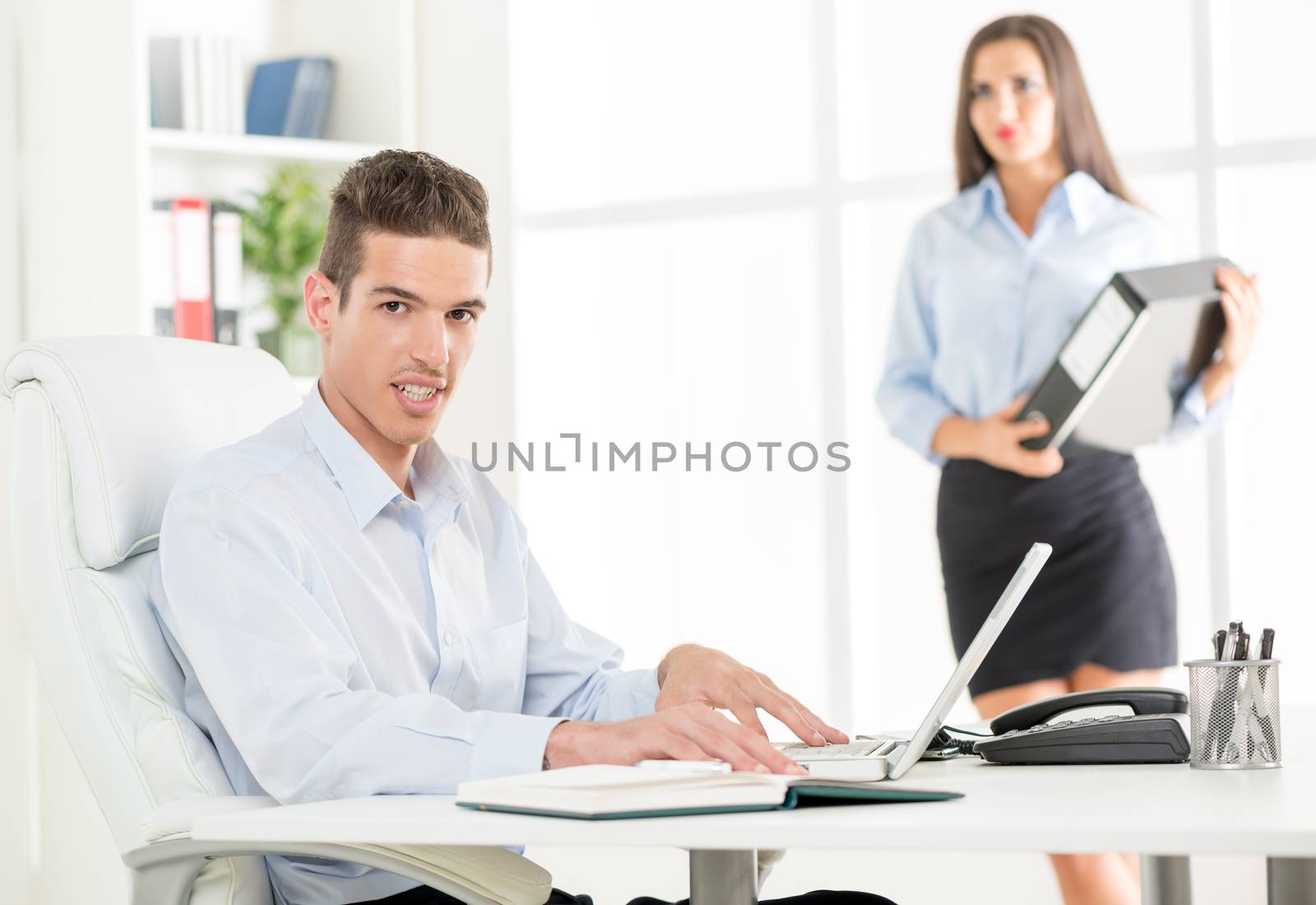 Young businessman sitting in office chair in front of a laptop, with a smile looking at the camera in the background is a young woman holding a binder.