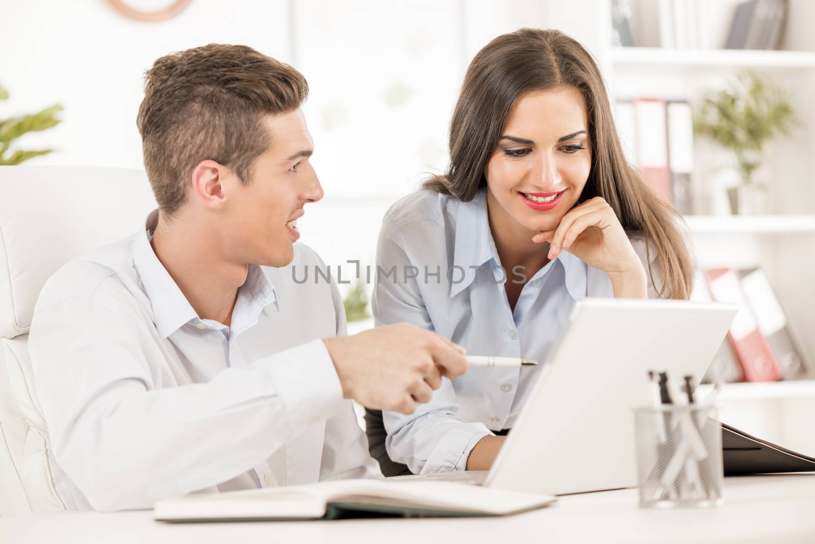 Young business people, businessman sitting at an office desk and showing something on a laptop, next to him is a businesswoman leaning against the table and looks at a laptop with a smile.