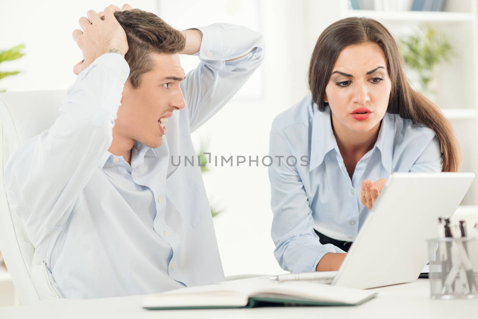 Two young concerned business people in the office, young businessman holding his head with a worried expression on his face looking at business woman who stands beside him, leaning on office desk with a sad expression on her face looking at  laptop.