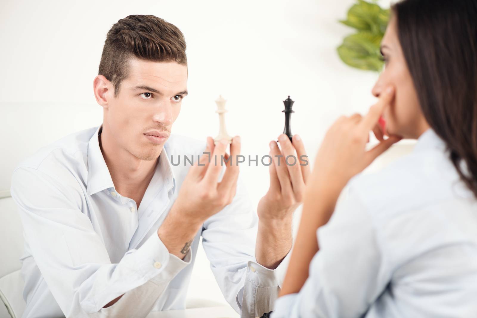 Two young people sitting at a table facing each other, young man holding a black and white chess figure with a serious expression on his face looking at the young woman who sits across from him.