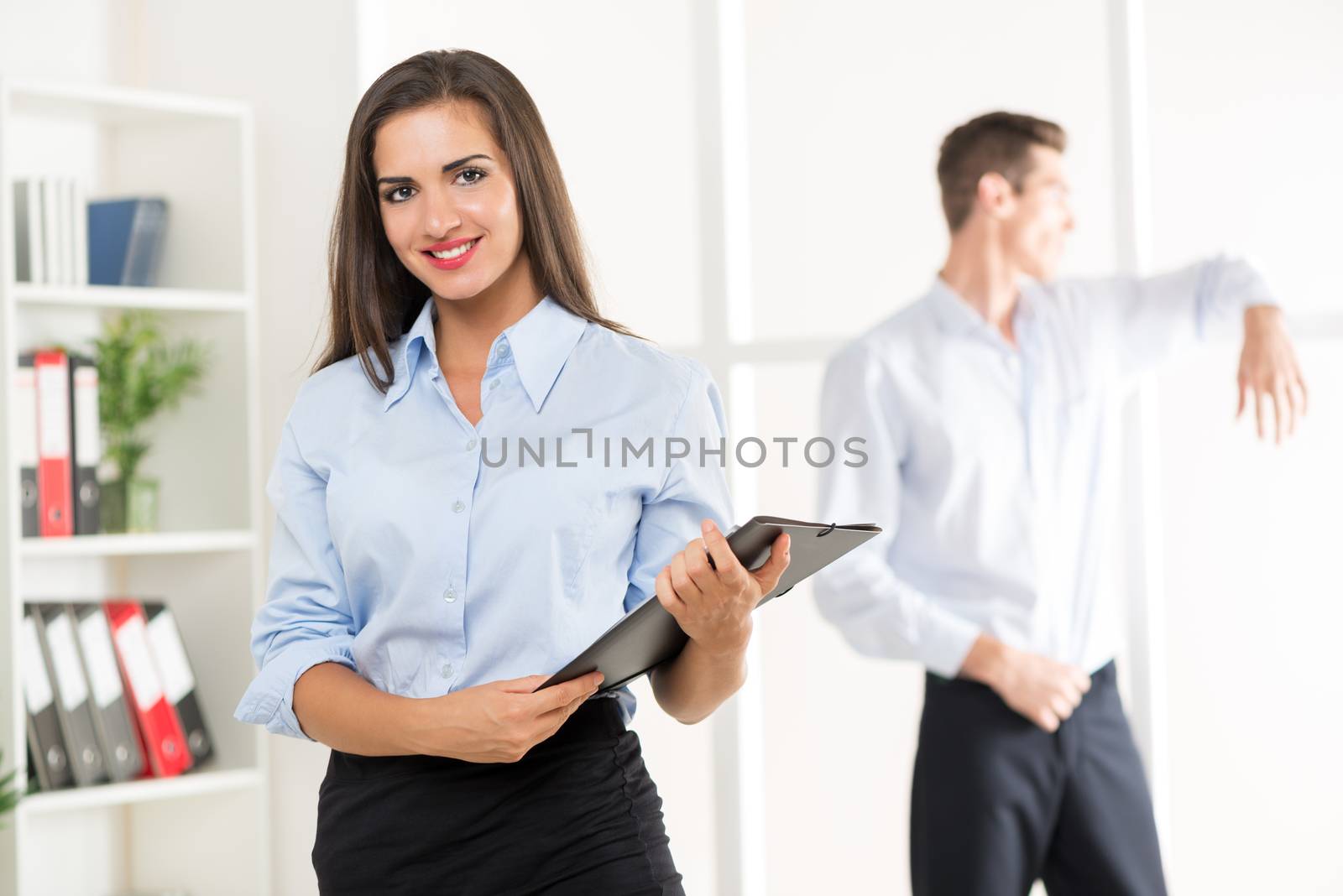 Young businesswoman standing in office with folder in hand, with a smile looking at the camera behind it is seen young businessman looking through the window of the office.