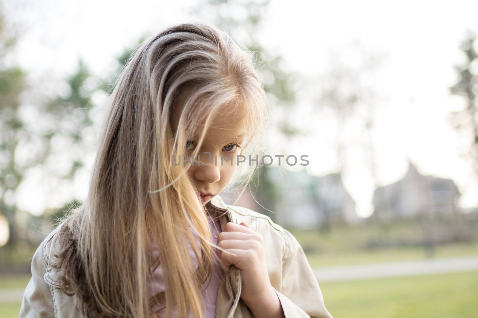 Close-up of a little girl with long hair standing alone outside with a sad expression on her face looking at the camera.