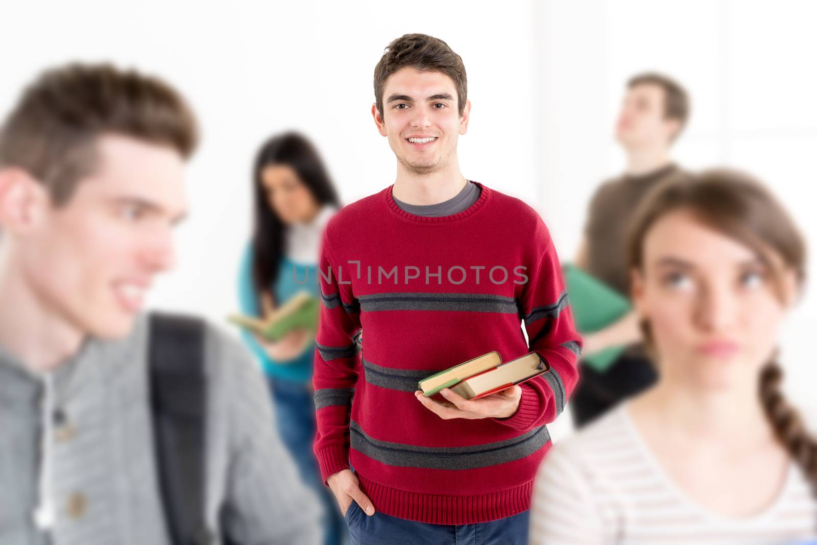 A beautiful smiling young man with books is standing in the foreground. A happy group of his friends is around him. Looking at camera.