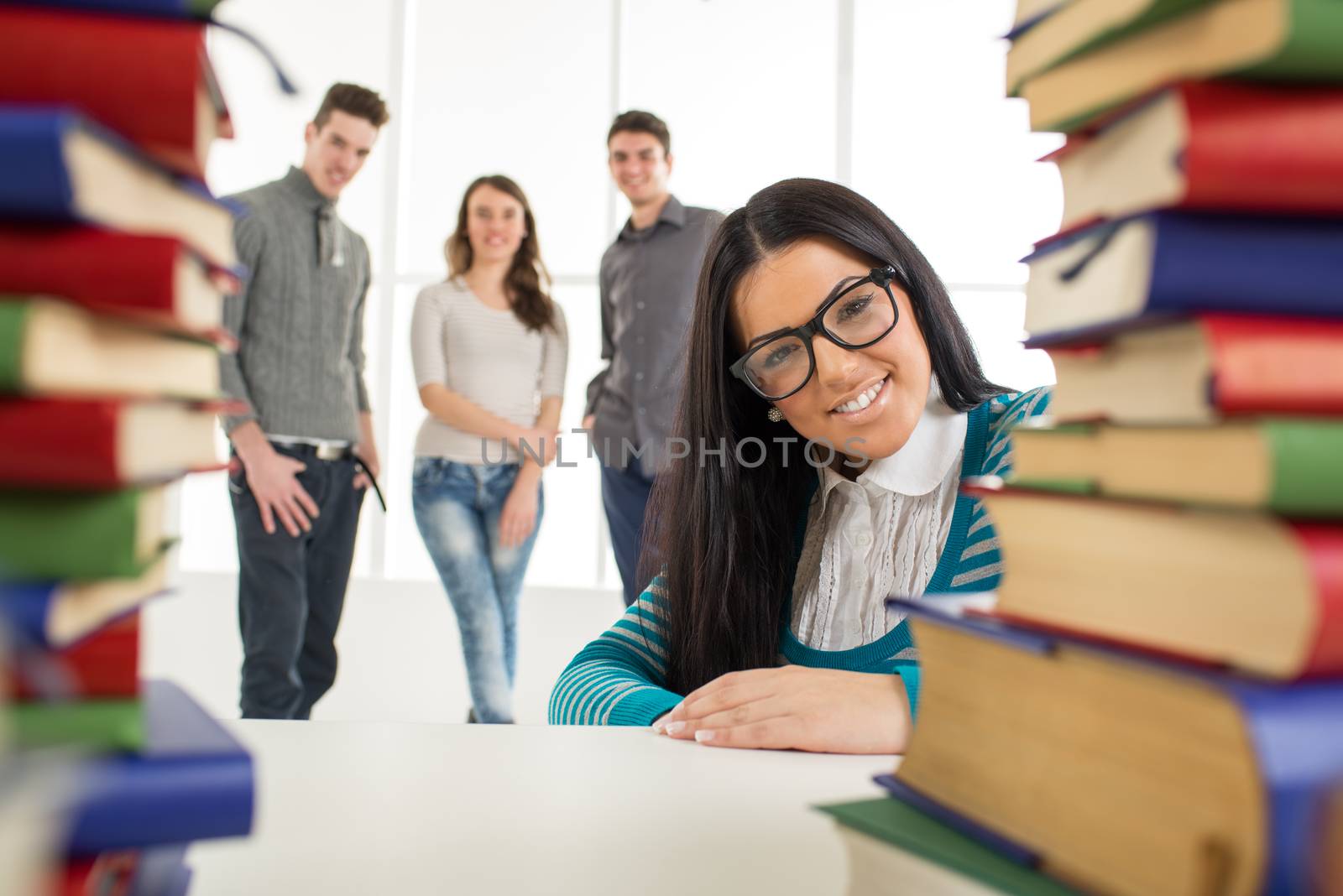 Portrait of a beautiful smiling girl with glasses sitting behind the many books in the foreground. Looking at camera.