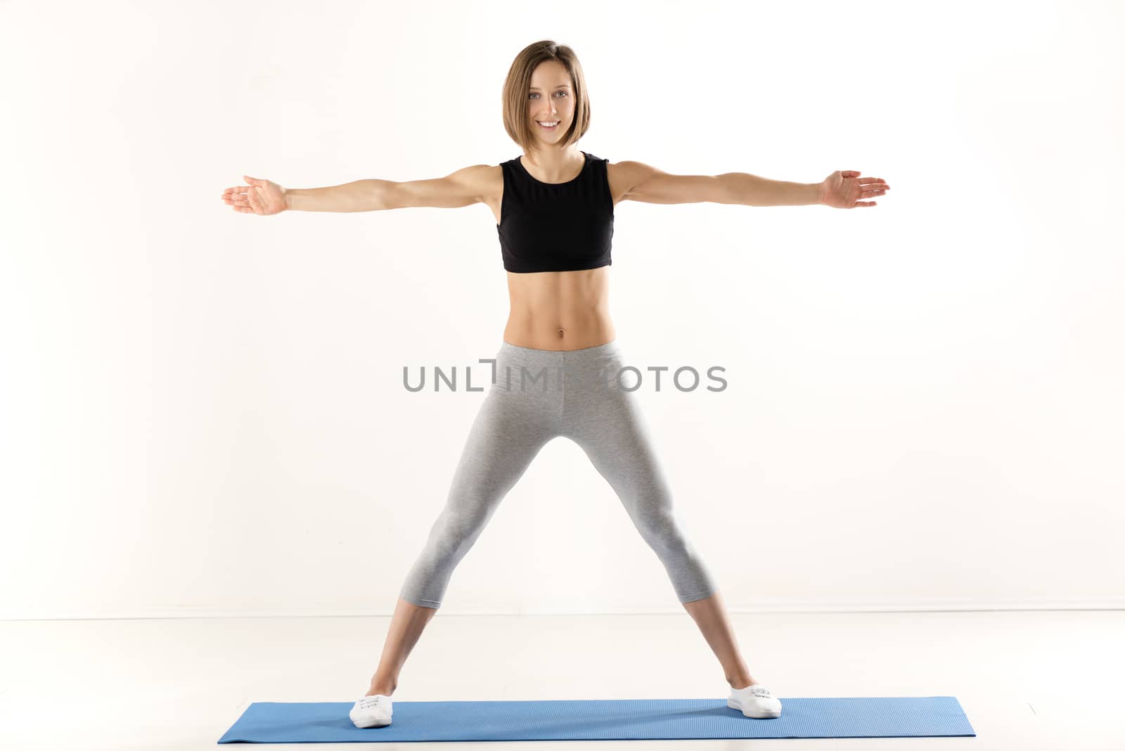 Athletic build women in sports clothes with arms outstretched, standing on the mat for exercise and with a smile looking at the camera.
