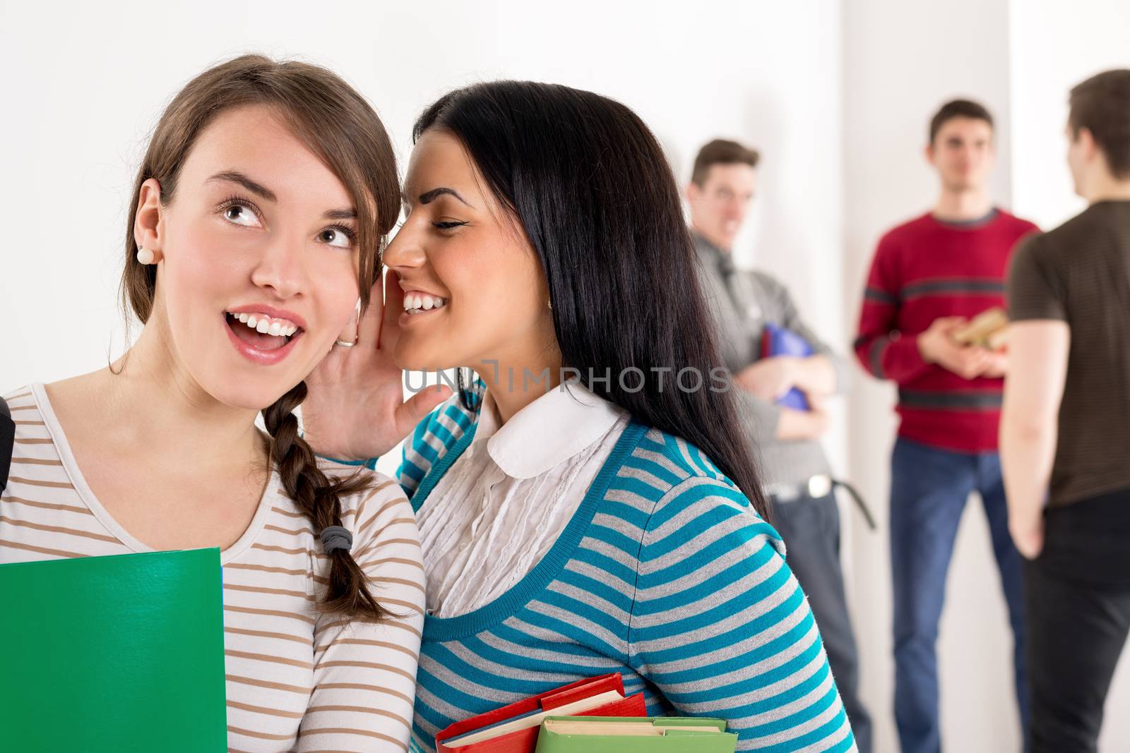 A beautiful teenage girl whispering with her cute friend. They is standing in the foreground. A group of men student is behind them and talking.