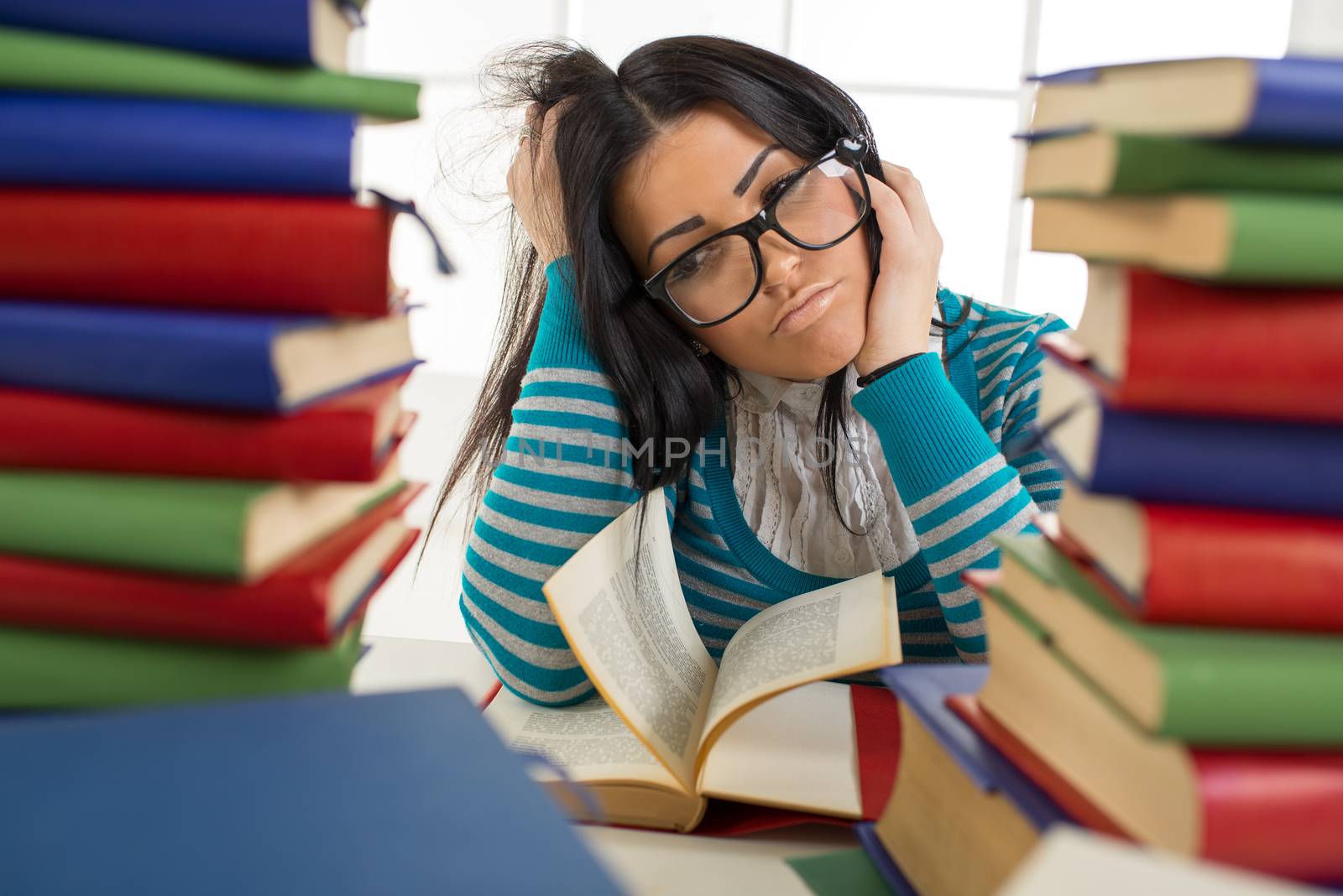 Portrait of tired student girl with glasses sitting and learning among the many books in the foreground. 