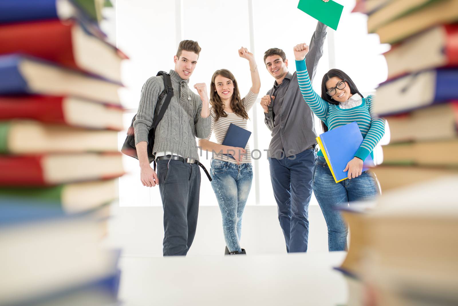 Group of cheerful students standing among the books with arms raised in a fist in school hall and looking at camera.